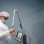 Médico utiliza equipamento de laser em procedimento dermatológico, usando proteção ocular, em ambiente clínico.