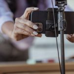 Pessoa posiciona um celular em um tripé para gravar um vídeo em ambiente interno.