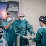 Equipe de profissionais de saúde se prepara para uma cirurgia em uma sala de operações, usando roupas e equipamentos de proteção.